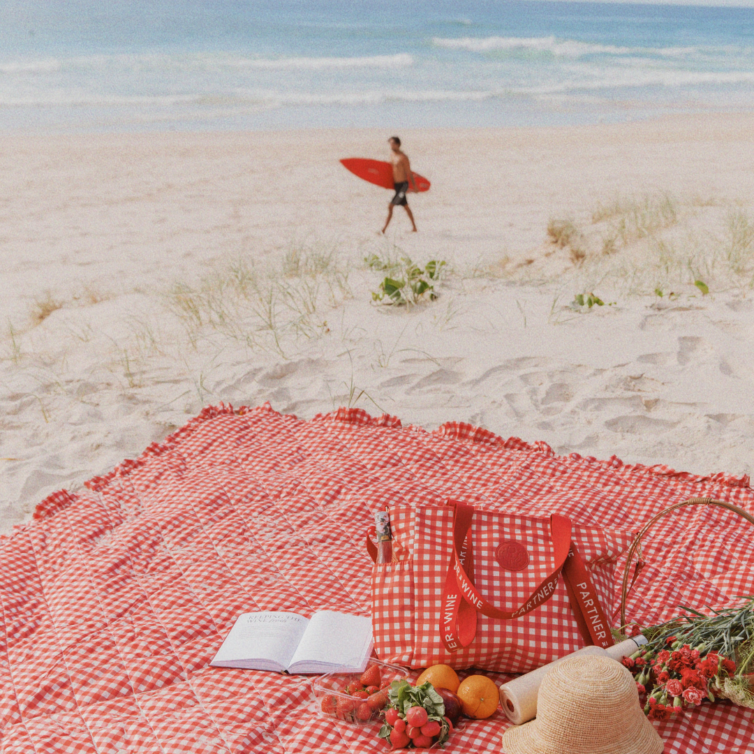 Person with a surfboard walking on a beach with a red gingham waterproof picnic blanket and picnic setup.